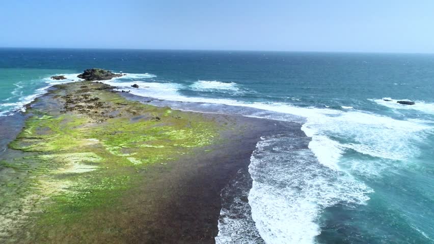 Aerial drone view of a small rocky island with green moss and waves crashing on a tropical coastline under a clear blue sky. Scenic seascape from above.