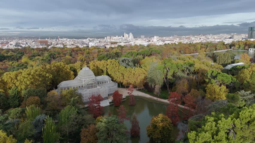 Aerial view of Palacio Cristal surrounded by autumn foliage and a serene lake, reflecting the cloudy sky, Madrid, Community of Madrid, Spain.