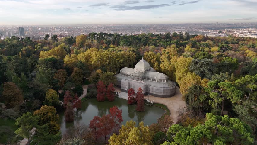 Aerial view of Palacio Cristal surrounded by trees with autumn colors and a pond reflecting its image in a tranquil scene, Madrid, Community of Madrid, Spain.