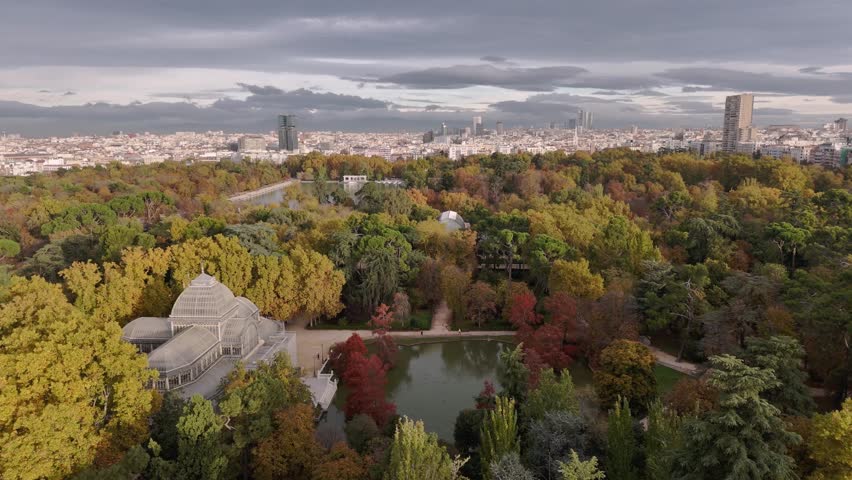 Aerial view of the Palacio Cristal surrounded by trees in autumn colors, contrasting with the grey city skyline and cloudy sky, Madrid, Community of Madrid, Spain.