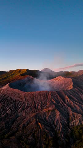 Vertical pull-back drone shot captures Mount Bromo’s smoking crater with Mount Semeru in the background, bathed in warm sunrise light over East Java’s volcanic landscape.
