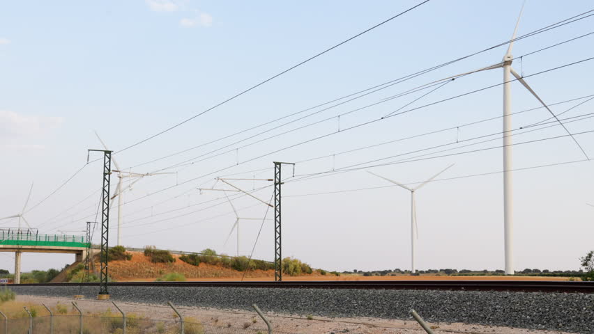 High-speed train passing in front of wind turbines in Motilla del Palancar, Spain, with power lines and railway infrastructure under clear summer sky