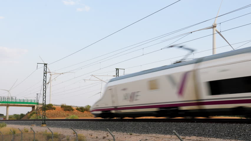 High-speed train passing in front of wind turbines in Motilla del Palancar, Spain, with power lines and railway infrastructure under clear summer sky