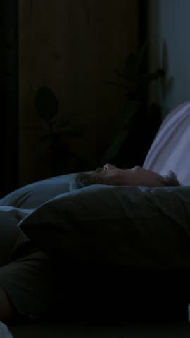 Vertical time lapse of Caucasian man with smartphone habit lying in bed at night, unable to sleep, turning and twisting, repeatedly checking social media for messages, face illuminated by blue light