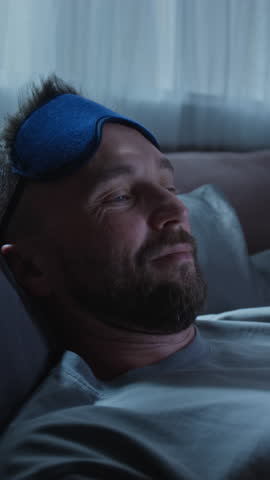 Vertical close-up of face of young Caucasian man checking electronic gadget at bedtime, browsing social media for messages, putting on eye mask and falling asleep