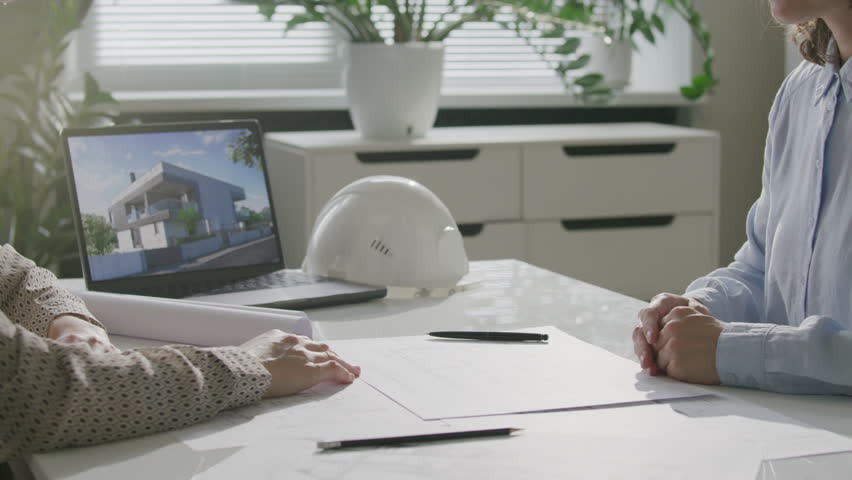 Two female architects shaking hands over desk with architectural plans and laptop, then discussing project during meeting in sunlit office. Close-up view