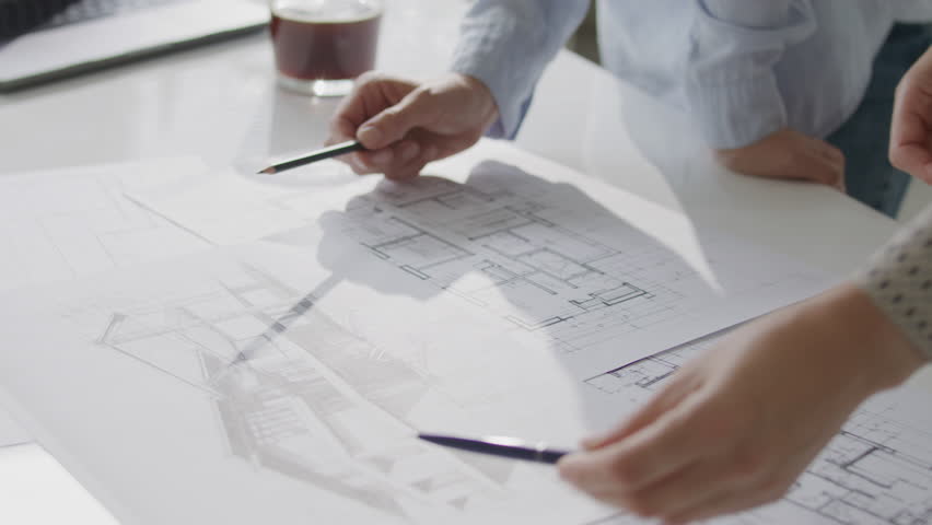 Close up view of hands of two female architects holding pencils while discussing technical building sketches and floor plans on office desk