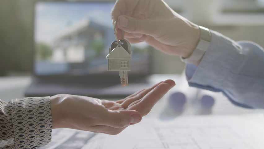 Close up view of hand of female property buyer receiving house shaped keychain with keys from seller over office desk during real estate handover
