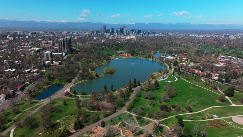 Leo City Park Denver Colorado Lake Pavilion vibrant spring summer aerial drone front range cityscape green lush grass trees blossom sunny blue sky snow capped Rocky Mountains circle left motion