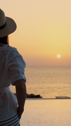 back view of young woman in summer clothes and sun hat, standing at the pool, enjoying the sunrise. summer. seaside resort. travel concept