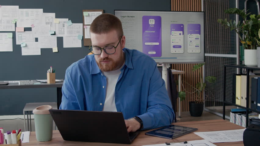 Portrait of young male UX designer typing on laptop creating UI prototype for mobile app then turning to camera with smile while working at office, slow motion