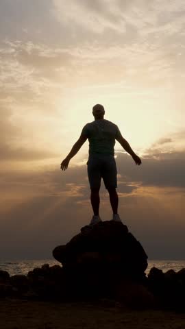 Dark male silhouette, man stands on rocks in front of the ocean or sea, with his arms outstretched and hands up at dawn, sunrise. concept of human strength and faith