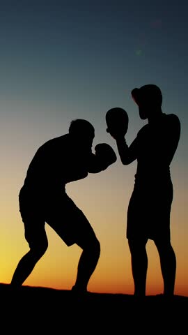 Two dark male figures, at sunrise, against the light, boxing, fighting in sparring, training in a pair of techniques of strikes, near water, in summer