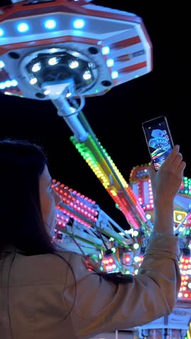 Woman hands are holding smartphone and taking photo or shooting video. Carousel with lights. Incredible colorful flashing light of vintage carousel. Popular Chair Swing Rides in Amusement Park