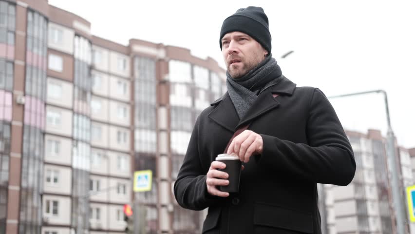 Handsome man wearing black coat and brown leather bag walking down on the city, drinking coffee paper takeaway cup with mobile phone. Stylish bearded middle-aged male using smartphone at urban street