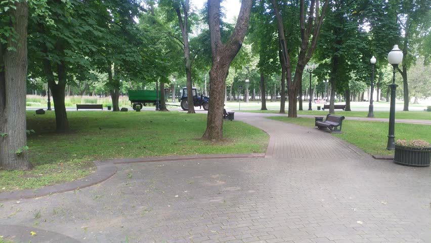 In the city park, trees grow on grassy lawns and there are tiled footpaths with benches and urns. A tractor with a bucket and trailer drives among them to clean up the area. Sunny summer weather