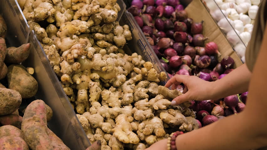 In a grocery market, a woman carefully chooses fresh ginger root among an array of organic vegan produce. Her hands sift through the vibrant display, slow motion