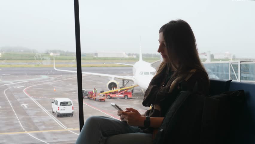 A woman using a smartphone while waiting for her flight at the airport against the background of an airplane