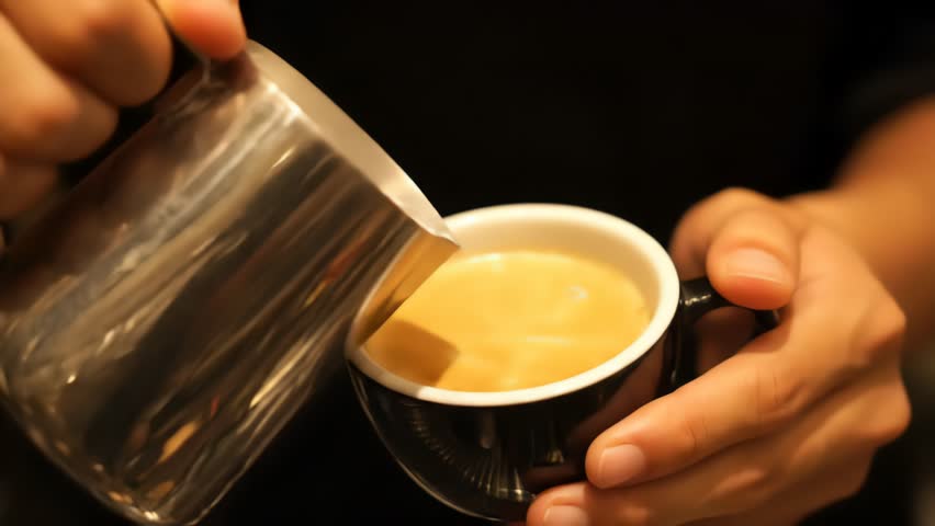 Barista pouring cream into a coffee cup creating latte art. The cup is black with a white latte art design. Close up shot.