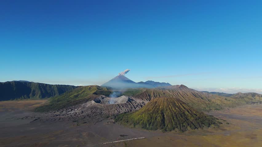 Forward drone shot captures Mount Semeru erupting above the Bromo Tengger caldera, ash plume rising behind steaming Bromo and ribbed Batok under clear East Java sky.
