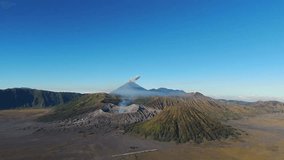 Forward drone shot captures Mount Semeru erupting above the Bromo Tengger caldera, ash plume rising behind steaming Bromo and ribbed Batok under clear East Java sky. - Powered by Shutterstock - Get 15% off with code: PIKWIZARD15