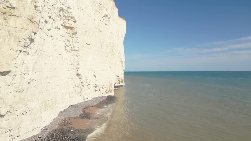 Dolly-out shot along the shoreline, revealing the towering white cliffs of the Seven Sisters meeting the calm blue waters.