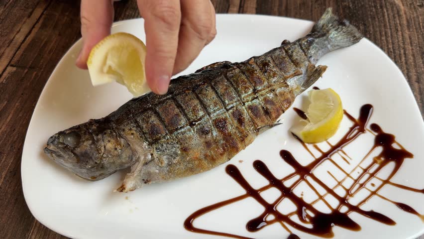 Whole river trout grilled in grill grate over a charcoal on a plate on a wooden table, view during pouring with juice from a squeezed lemon
