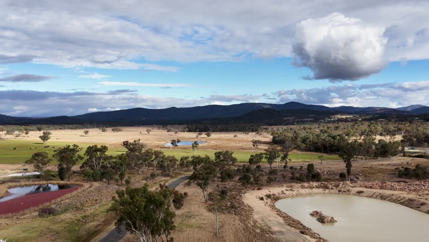 Drone camera smoothly pans across a rural Australian landscape with farm ponds, open fields, scattered trees, and distant hills under partly cloudy daylight