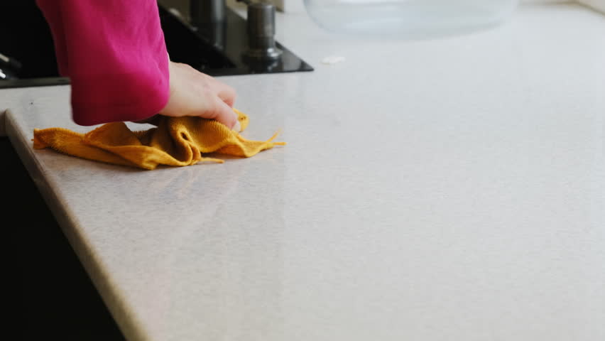 Woman presses yellow cloth against kitchen counter removing visible dirt. Fingers grip fabric firmly creating swift circular motion