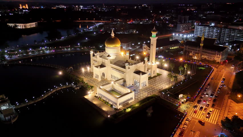 Rotating aerial drone shot orbiting the Omar Ali Saifuddien Mosque lit up by it's lights at nighttime in Brunei. Epic cinematic aerial rotating drone shot.