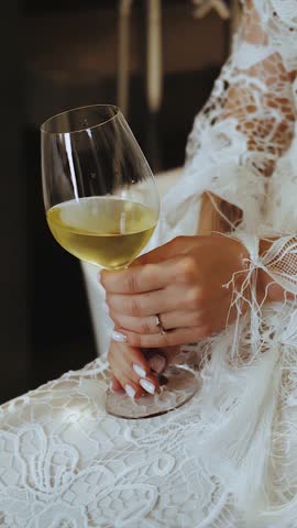 Vertical video. Bride in boho style dress holding a glass of wine while sitting on a chair, close-up of hands. Woman relaxing with a glass of wine. Slow motion.