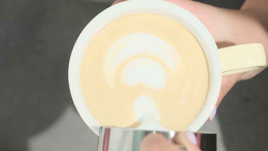 Woman gently pours milk into coffee resulting in picture on top. Barista professionally shows last step of creating latte. Process of making coffee