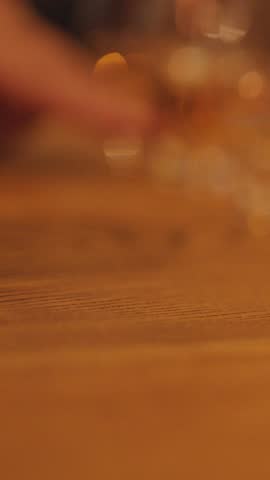 A hand gently slides a whisky glass across a rustic wooden table in soft, ambient lighting. Shallow depth of field and subtle motion blur create a cozy, intimate mood
