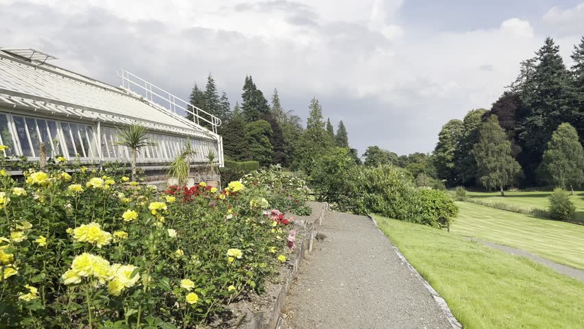 Panning across to a historic ornamental greenhouse full of plants. Outside the greenhouse is a heavily planted bed featuring mostly yellow roses.
