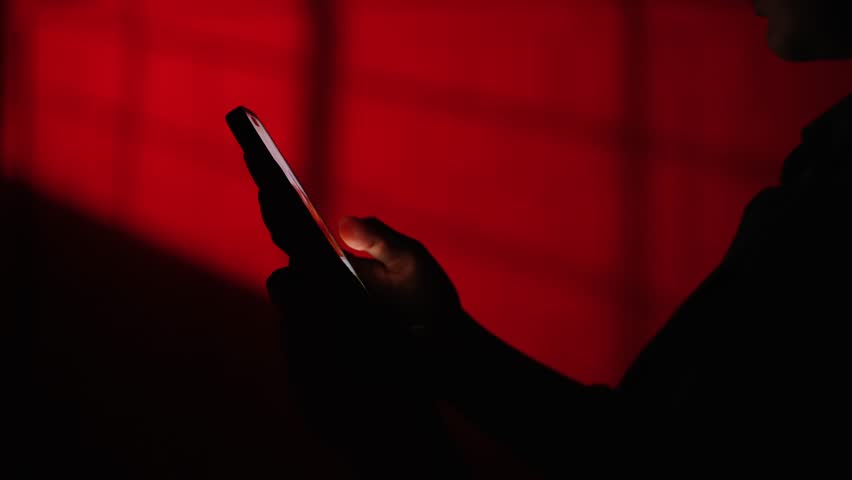 Shadowed figure operates a smartphone against a striking red backdrop, hinting at themes of technology and communication.