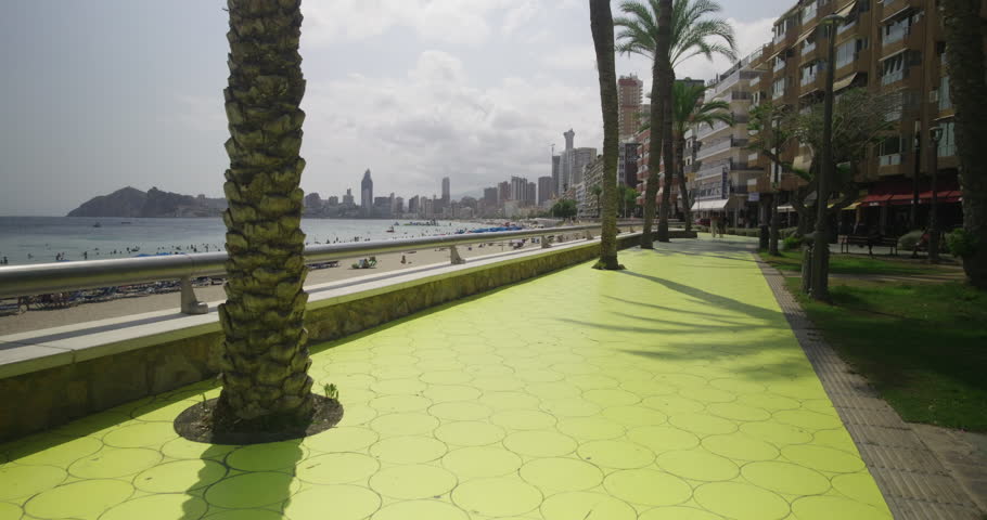 A sunny promenade with palm trees overlooks the sandy beach and skyline of Benidorm, Spain. Tourists enjoy the seaside atmosphere with modern buildings rising in the background.
