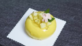 Small yellow mango mousse cake decorated with gold leaf, diced mango, pink flower, and green leaves, sitting on a white doily on a dark gray surface, creating a visually appealing gourmet dessert - Powered by Shutterstock - Get 15% off with code: PIKWIZARD15