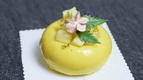 Small round mango mousse cake decorated with pineapple chunks, edible gold leaf, pink icing flower and fresh green leaves, sitting on white doily on dark gray surface - Powered by Shutterstock - Get 15% off with code: PIKWIZARD15
