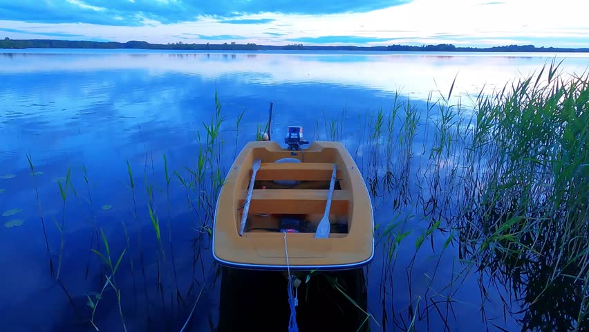 A small motorboat is docked on the shoreline of the Swedish lake in the countryside. The boat is orange-colored and has both a motor and oars for rowing. Calm lake view in the evening.