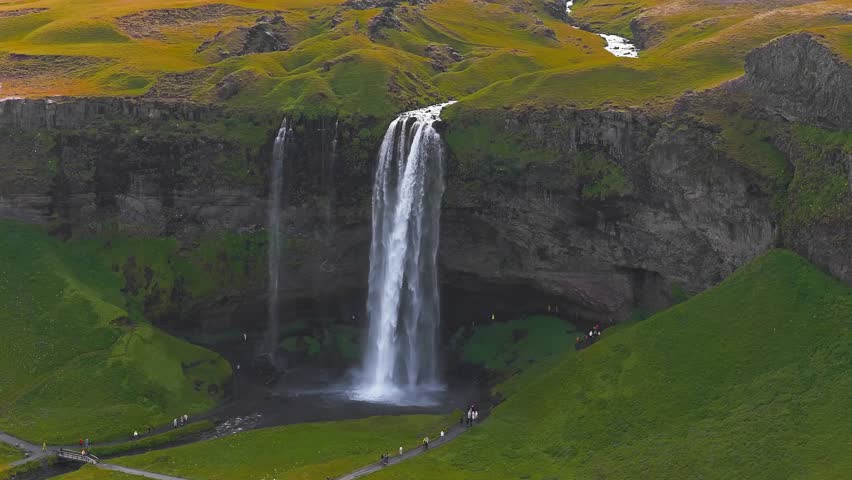 Seljalandsfoss waterfall cascades from a high cliff in Iceland, surrounded by green terrain. Visitors walk pathways, some venturing behind the falls.