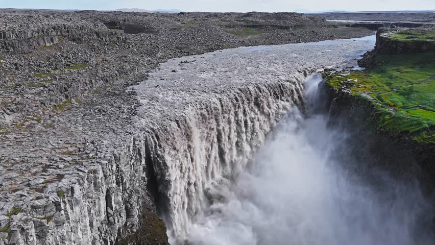 Aerial view of Dettifoss waterfall in Iceland, showcasing dynamic water motion, mist filled gorge, green moss, and rugged rocky terrain.