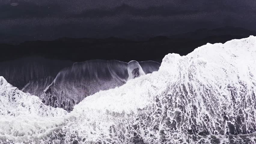 Aerial view of a black sand beach in Iceland as white waves crash and retreat, creating rhythmic motion and foamy patterns on the dark shoreline.