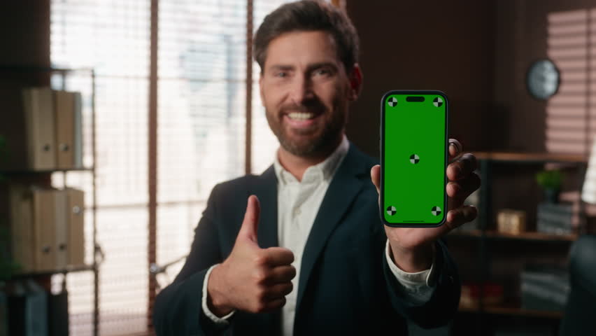 Smiling happy middle aged business man showing green screen mobile phone like gesture thumb up holding smartphone chroma key display success achieve mature businessman male looking at camera in office