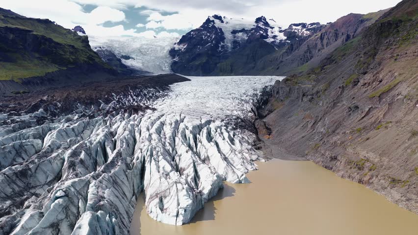 Sweeping aerial view of a massive glacier in Iceland, surrounded by rugged mountains, rocky cliffs, and snow capped peaks with visible crevices.