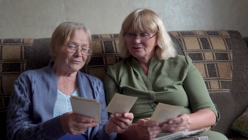 Old female mother with mature woman daughter sitting together on couch in house. Women reminisce while browsing through collection old retro photos. Family gatherings where sharing stories of past