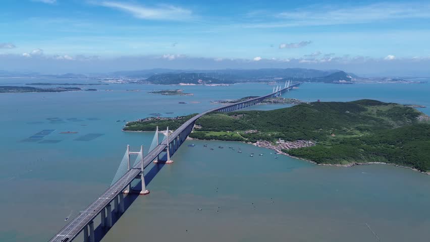 Aerial video of Pingtan Strait Rail-cum-Road Bridge on a sunny summer day, with its grand structure spanning turquoise waters, surrounded by lush green islands and clear blue skies.