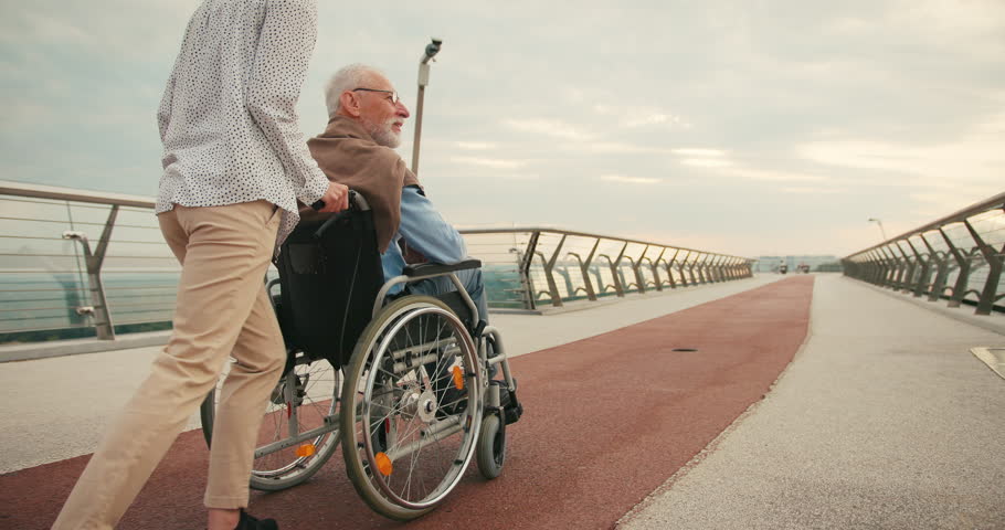 Young woman runs with elderly man in wheelchair happily stretching arms flying on bridge. Sense of unity and shared joy highlight strong connection