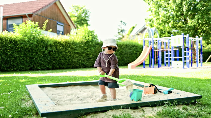 Stylish boy 2 years old in a T-shirt and shorts playing in the sandbox. Toddler in a Panama hat on his head and sunglasses digging sand with a plastic shovel. Child playing outside on the playground.