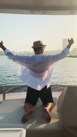 Handsome man with hat enjoying sunset sail on luxury yacht in Dubai Marina with hands wide open up in the air, vertical video