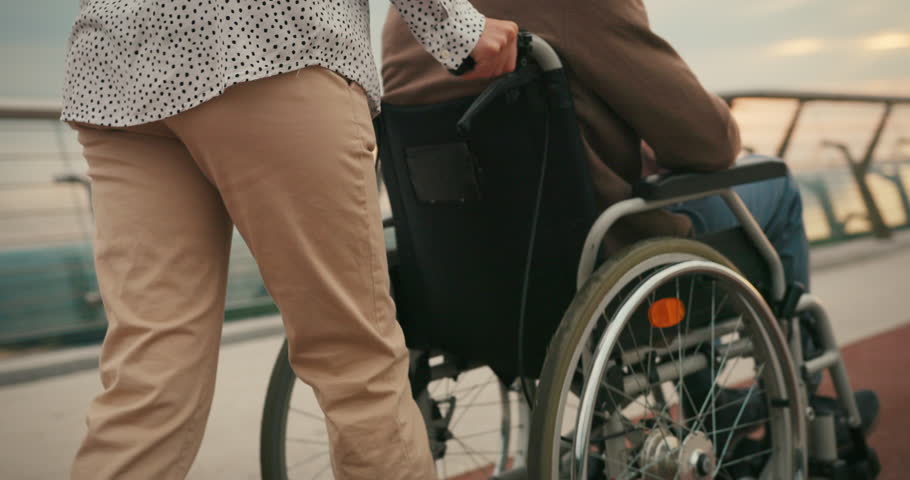 Woman pushes man in wheelchair on pedestrian bridge with hands resting in lap at sunset. Concept of support and enjoying peaceful moment together